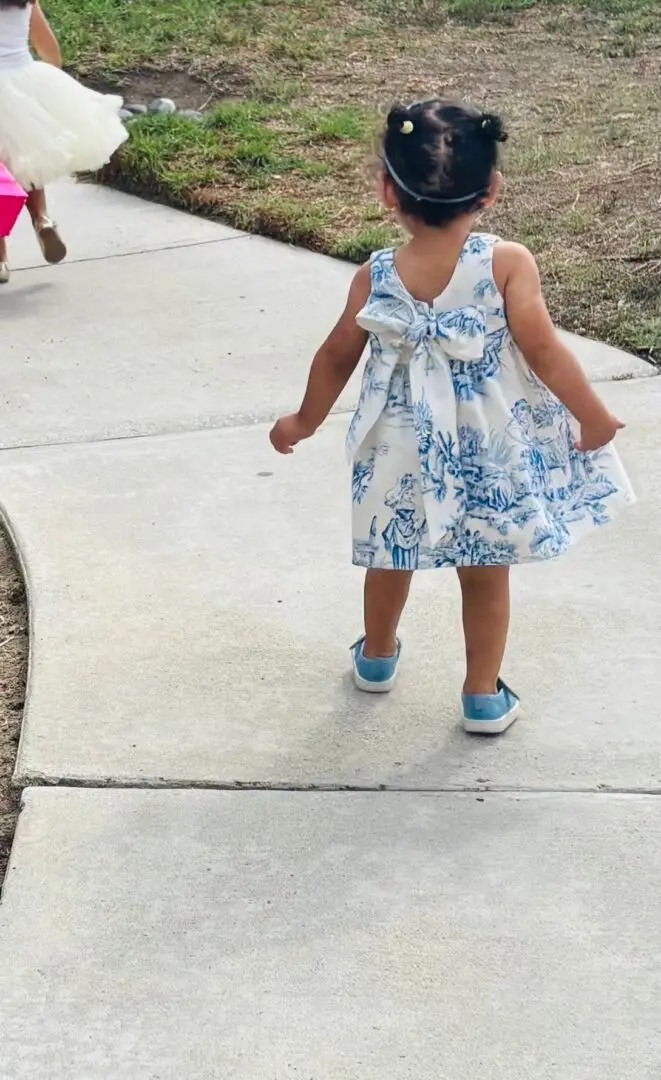 Toddler girl in a blue floral dress walking on a sidewalk.